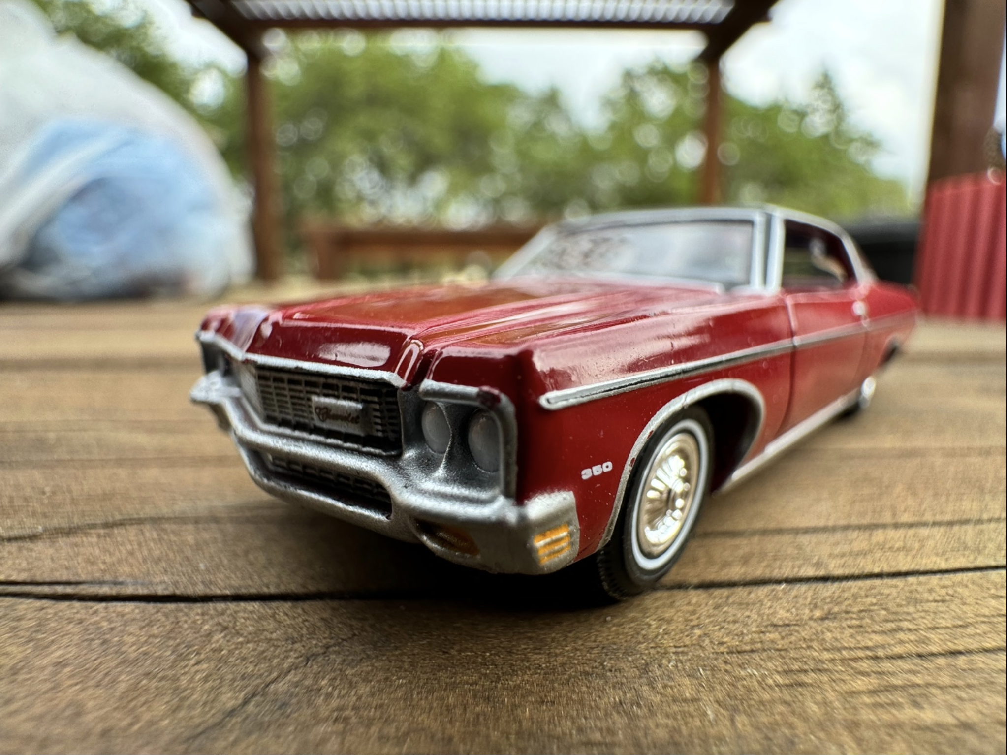 A close-up photography of a classic car toy model, specifically a 1970 Chevrolet Impala Coupe, on a wooden surface. The car is depicted with a glossy red paint, chrome detailing, and realistic styling. White-walled tires and silver hubcaps are visible, with clear windows and an intricate grille. The background is outdoor with a blurred bokeh effect featuring greenery, structures, and a blue sky. The image has a shallow depth of field, sharply focusing on the car while the background is out of focus.